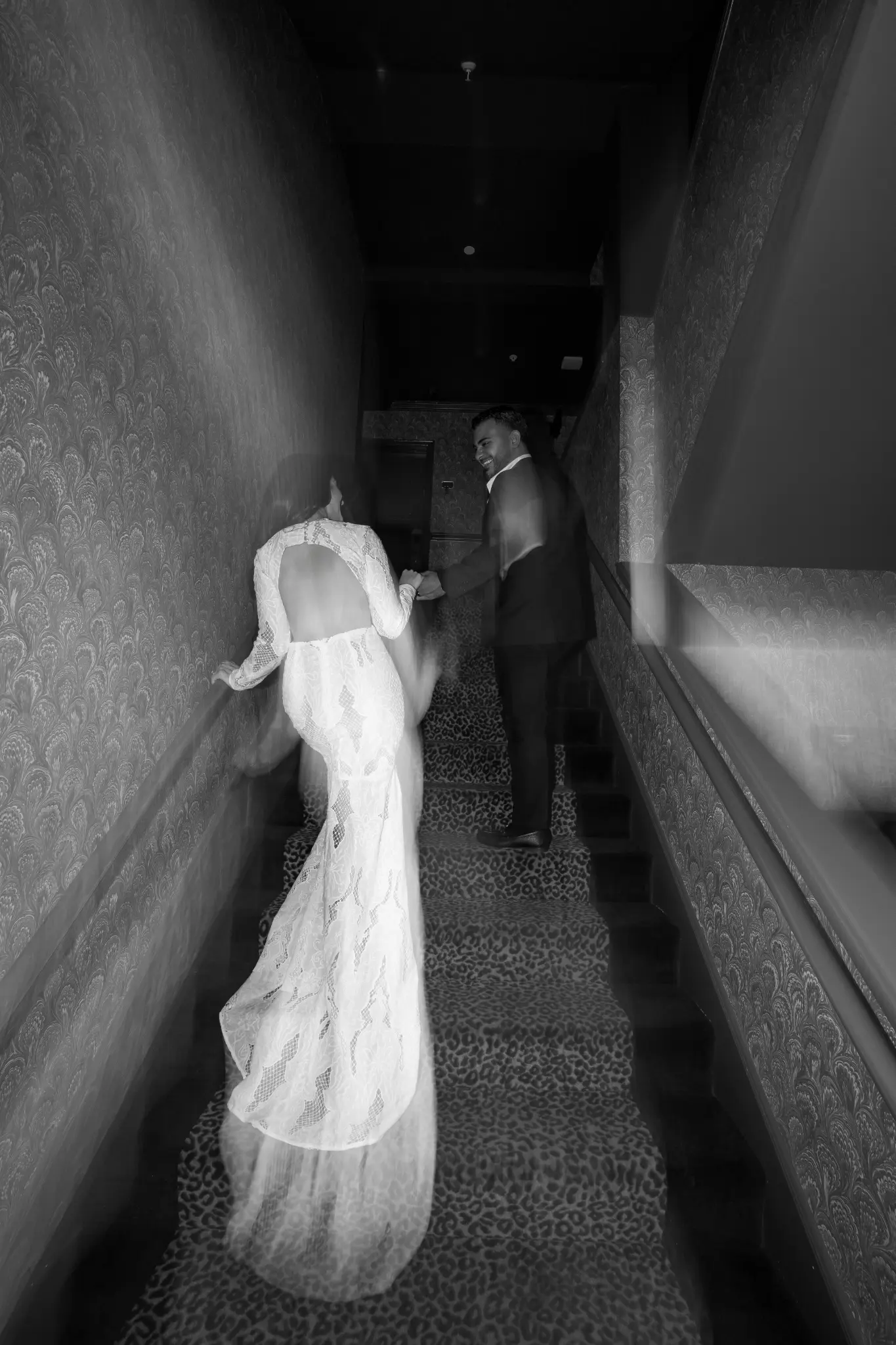 Black and white low-angle view of a couple in formal attire walking up a leopard-print staircase with motion blur