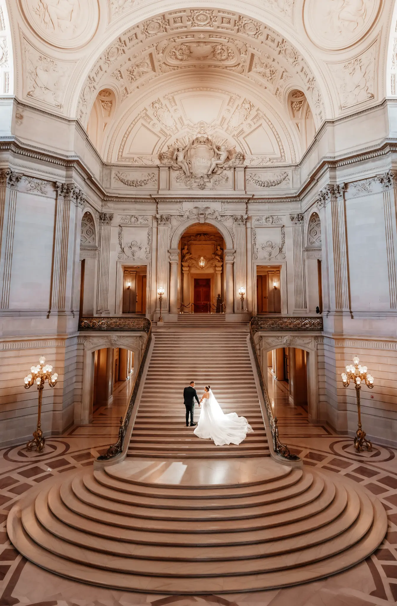 Couple in a tuxedo and white gown with a long train standing on a grand marble staircase in an ornate classical hall