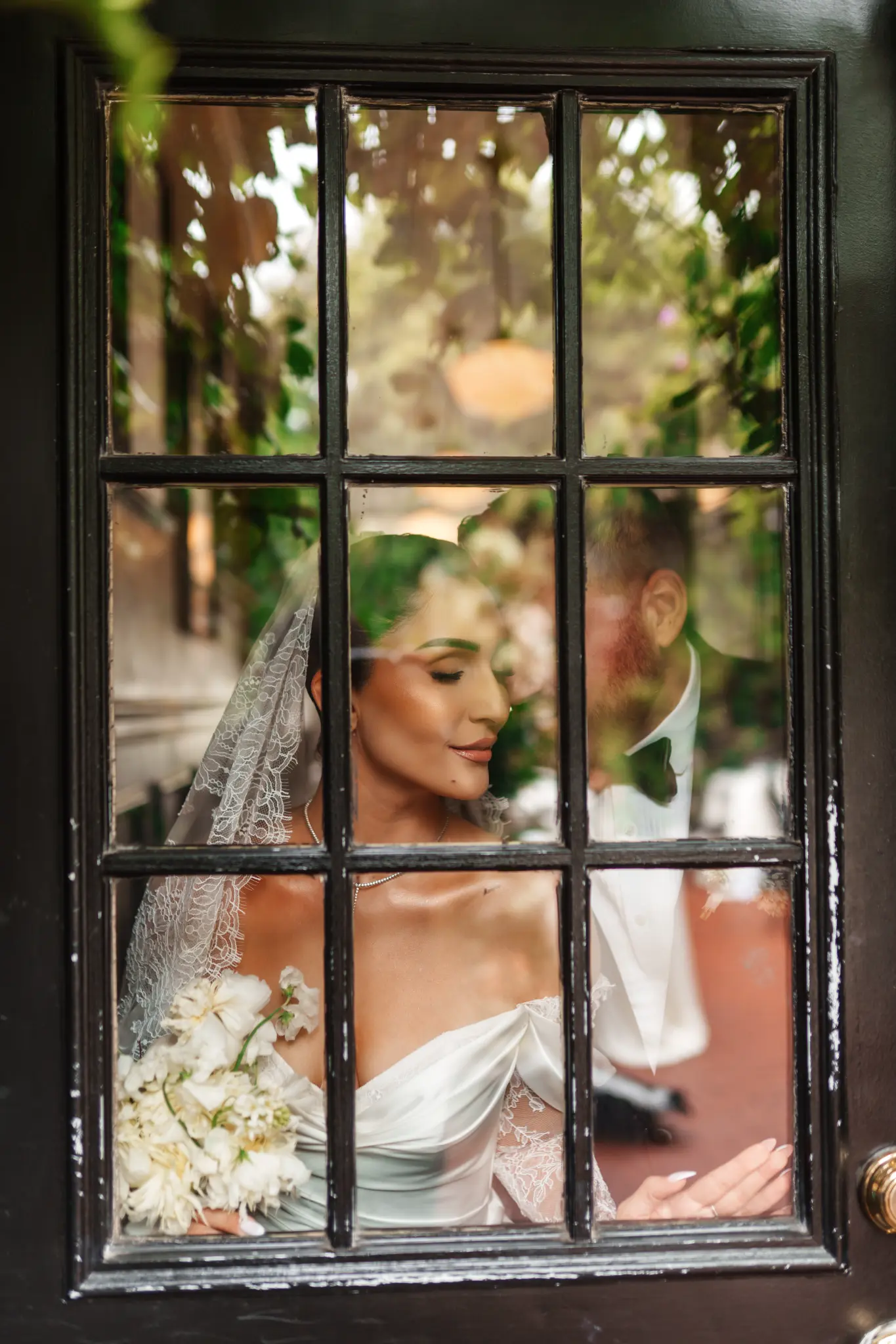 Bride in a lace veil looking down beside a glass door reflecting green foliage