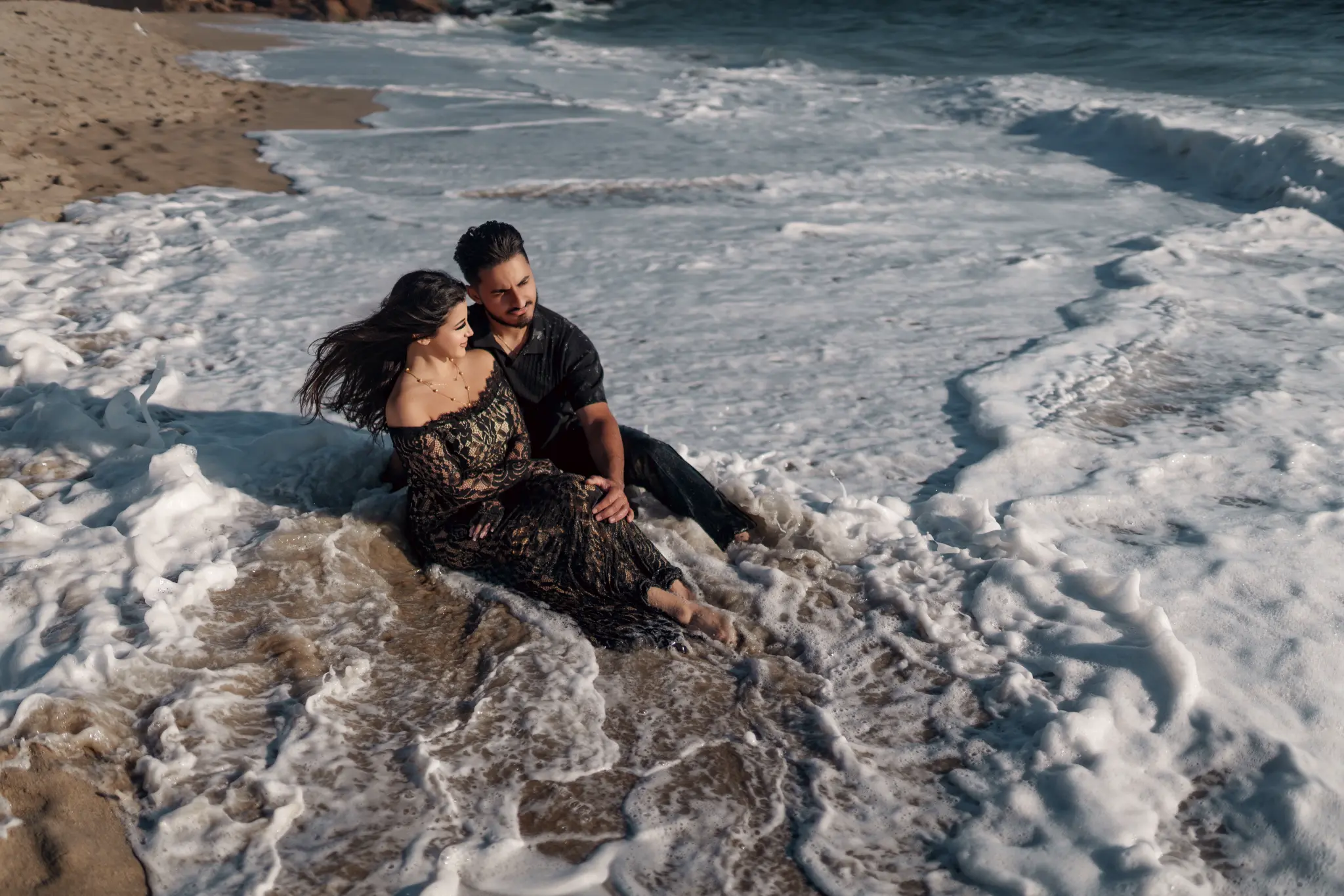 High-angle portrait of a couple sitting in sea foam on a sandy beach